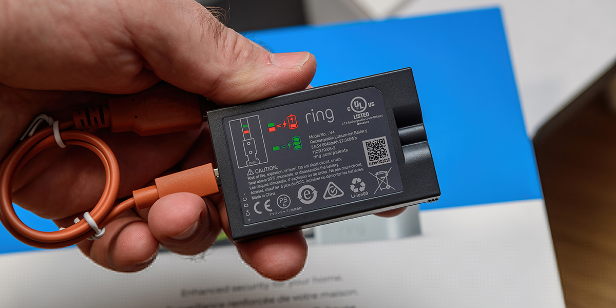 A hand holding a black smart Ring doorbell battery attached to an orange cord