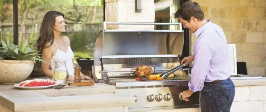 A man at a coyote grill set into a kitchen island. he is roasting vegetables and a chicken. A woman is on the other side of the island, smiling.