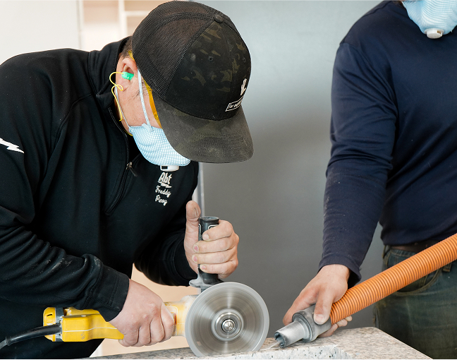 Technician wearing protective gear using a handheld circular saw to cut countertop material, with a second person assisting by vacuuming dust.