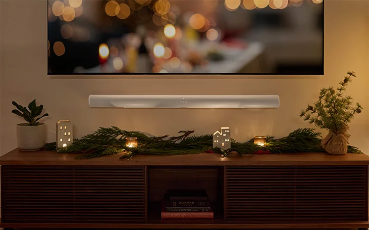 Sonos Arc Ultra mounted underneath a TV with dim lighting and holiday lights on the tv and holiday christmas garland on the entertainment center underneath the mounted soundbar and tv