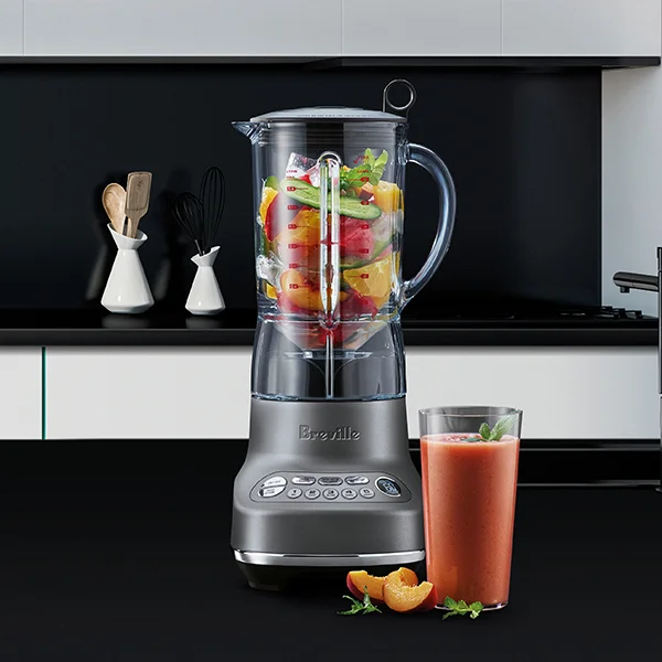 A silver Breville blender filled with fruit sitting on a black kitchen counter next to a pink smoothie