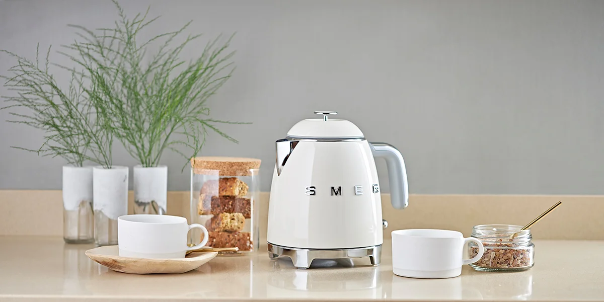 A white Smeg water kettle sitting on a countertop next to snacks, two tea cups and some plants