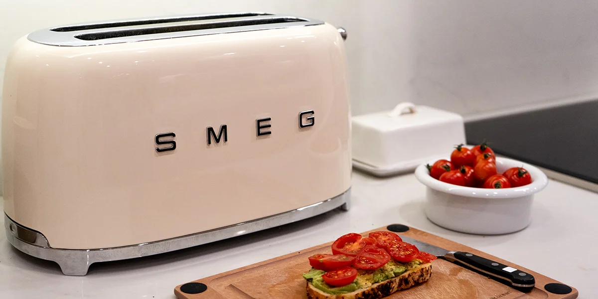 A smeg toaster on a kitchen counter beside a slice of avocado toast on a cutting board and some ripe cherry tomatoes
