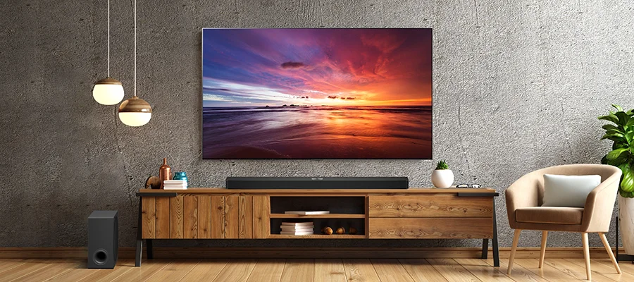 The LG S90TR soundbar in a grey room sitting on a TV stand beneath a screen that's displaying a colorful sunset