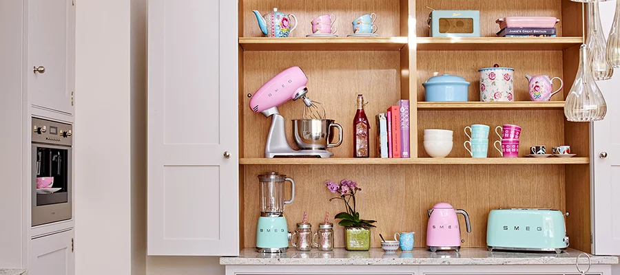 Pastel pink and blue small kitchen appliances sitting on kitchen shelves beside other kitchen tools, cookbooks and more.