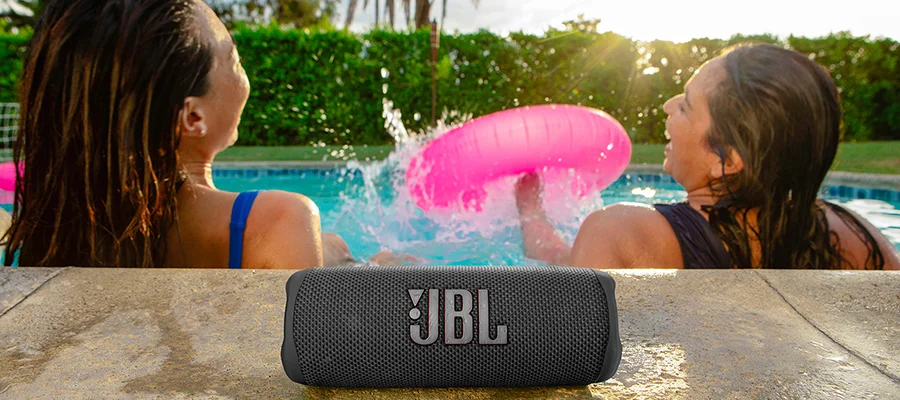 A black JBL Flip 6 speaker sitting on the pool deck behind two women