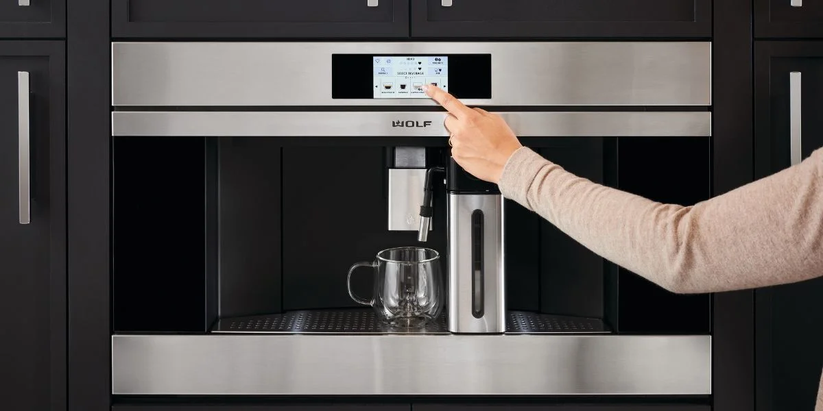 A stainless steel Wolf built-in coffee machine. The arm of a person offscreen is using the touchscreen to program their coffee choice.