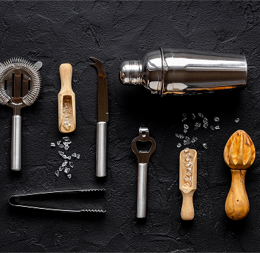 Overhead shot of a Cocktail shaker, cocktail strainer, ice tongs, bottle openeer and handheld juicer, showcasing the variety of bareware used to make a cocktail