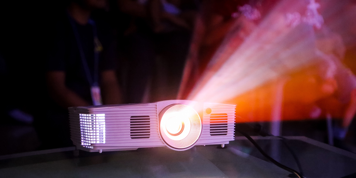 A white projector shining light and projecting an image with people sitting in the dark behind it