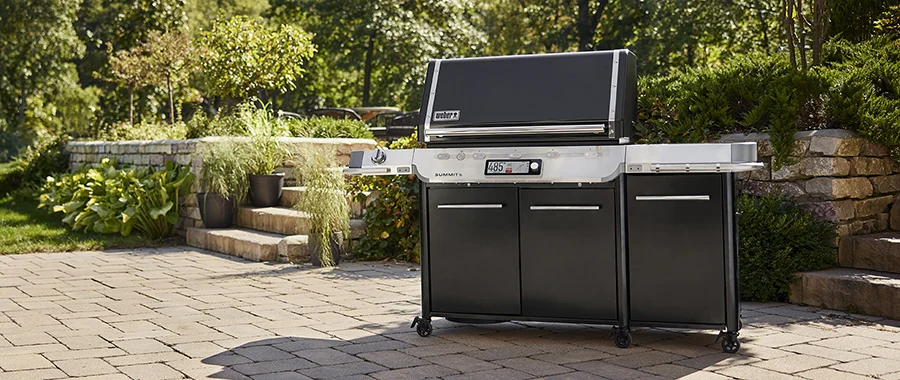 A black Weber Summit grill sitting outside on a stone patio near some greenery
