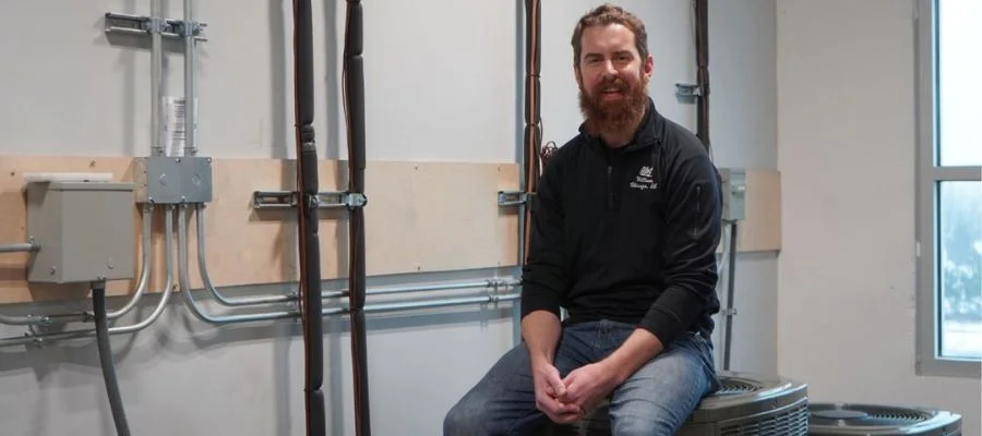 Bill O'Callaghan smiles and sits atop a central air conditioner in our HVAC training studio.
