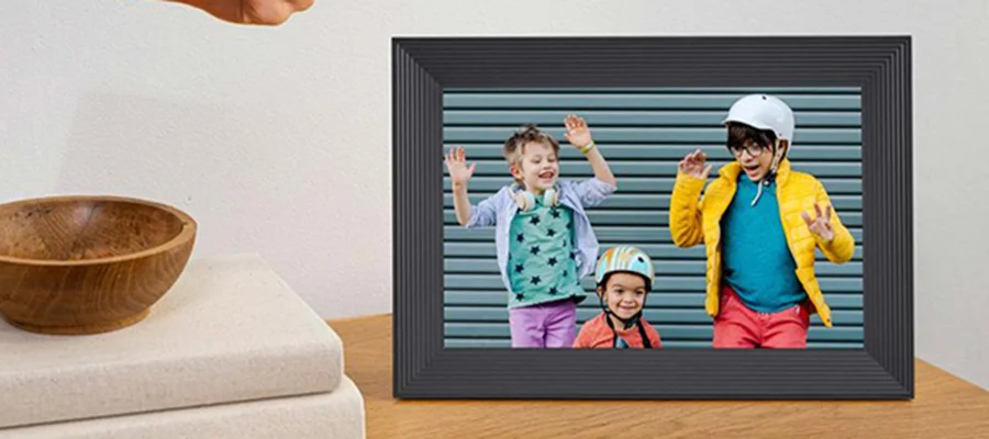 A hand holding an iPhone uploading photos onto the Aura Carver digital picture frame, which is displaying a picture of three children playing