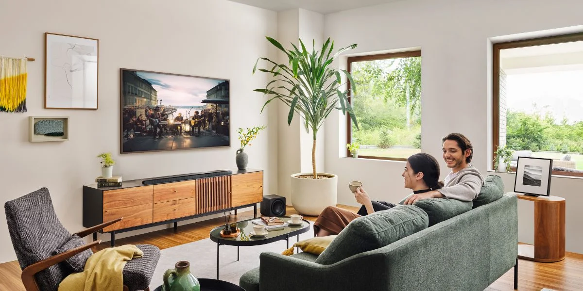 A couple in their living room on a green sofa watching TV. The room is brightly lit and the two have other framed art on the wall.