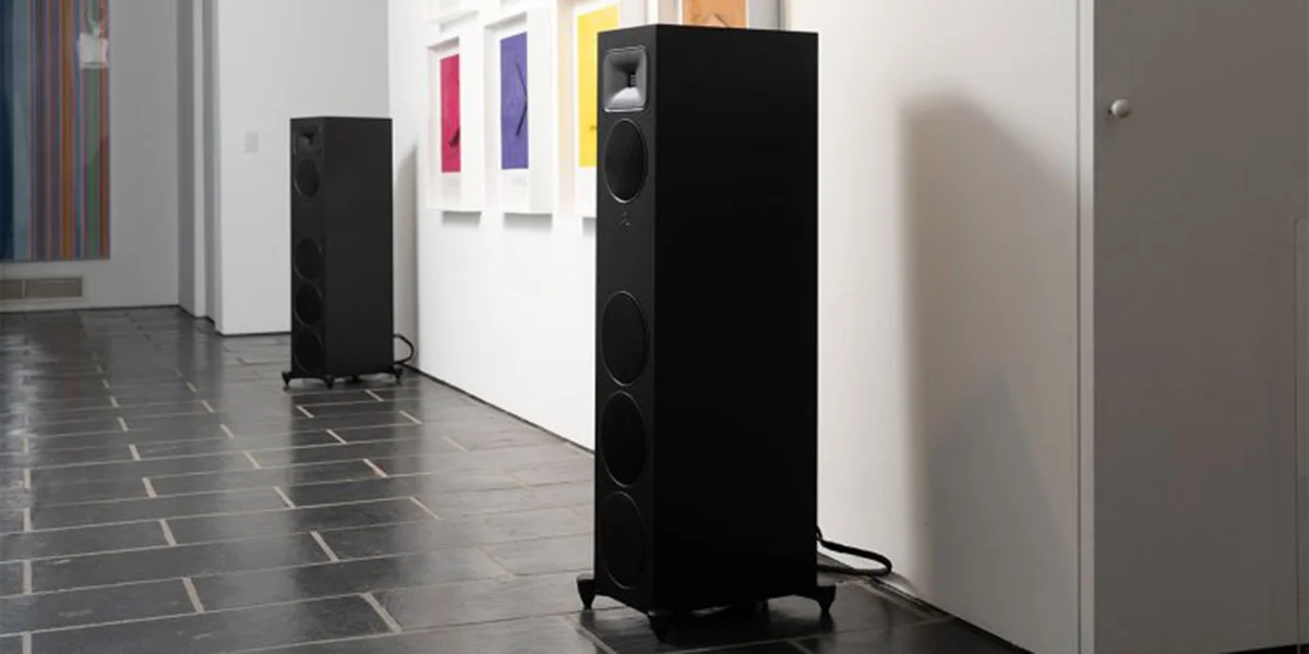 Two floorstanding speakers in a hallway. Between them is a white wall with solid colorful framed paintings.