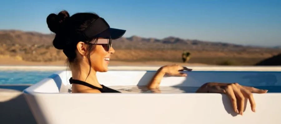A woman in a cold plunge wearing a black hat, sunglasses and smiling against the backdrop of mountains and water