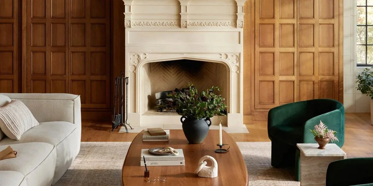 A large living room with wood walls centered around a white fireplace. Green armchairs and a white sofa sit on a large beige rug.