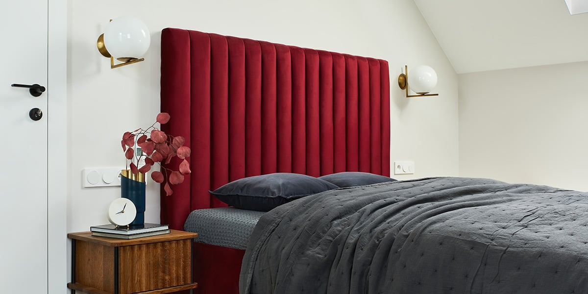 An image of a very small bedroom with a bed that takes up nearly the entire space. An extra-tall red headboard catches the eye, while lamps are built into the wall