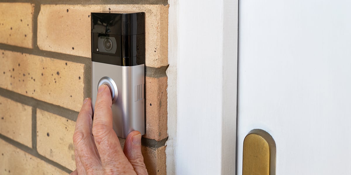 A finger pressing the button on a silver and black smart Ring doorbell outside of a home