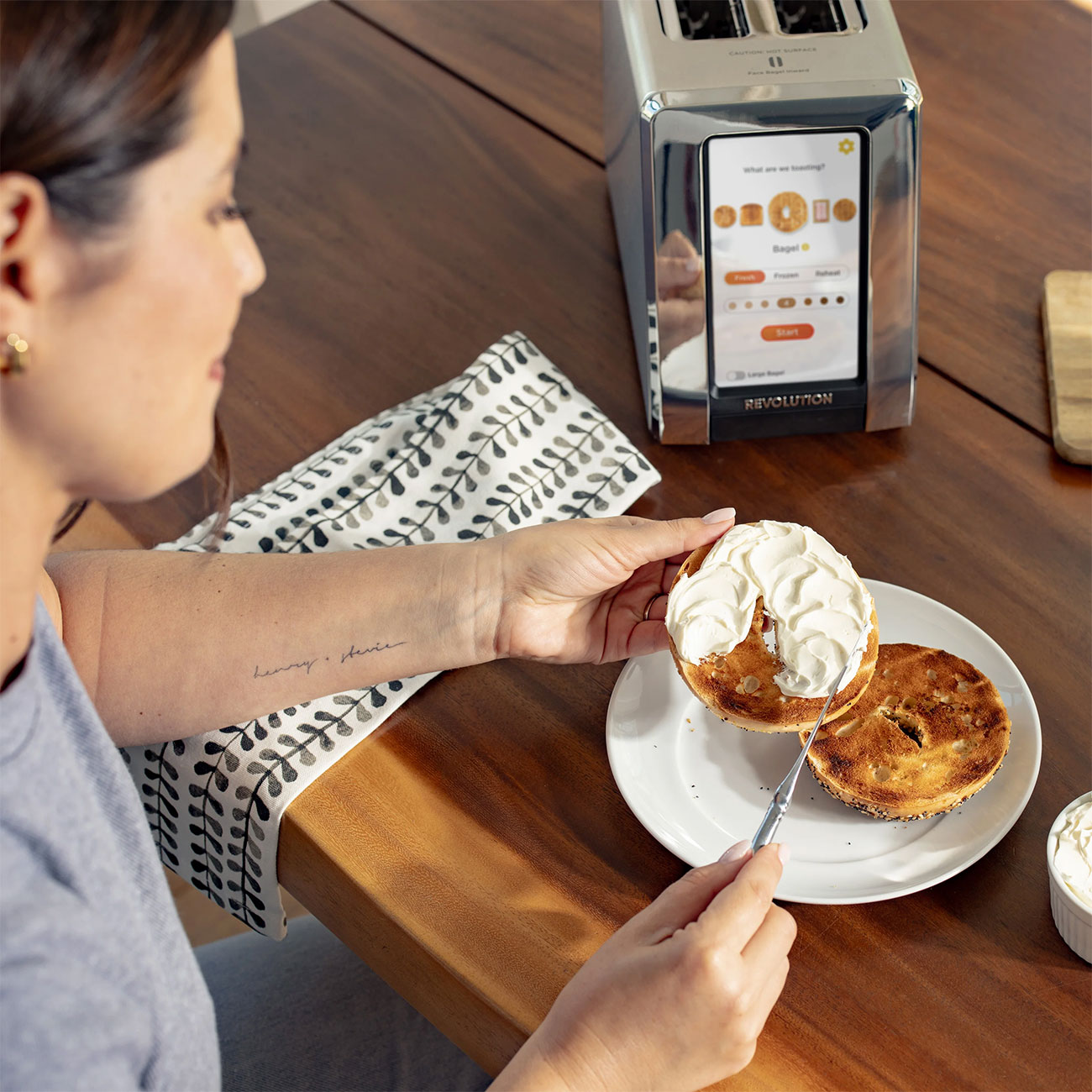 Woman Spreading Cream Cheese on Bagel with Revolution Cooking Toaster on Table