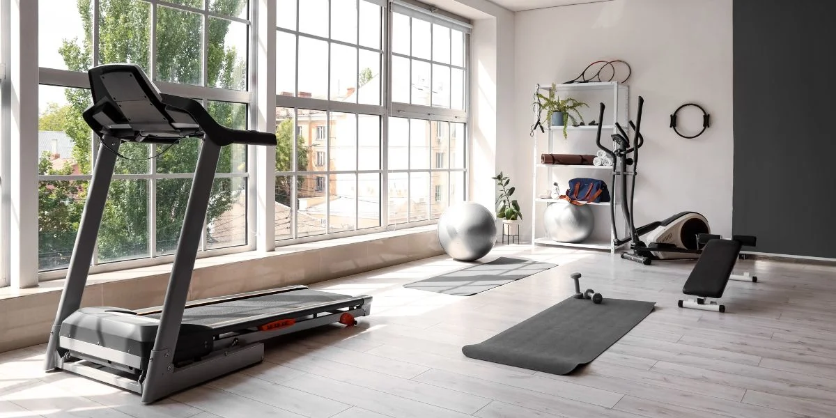 Modern home gym with treadmill, elliptical and weight bench in a room with large windows