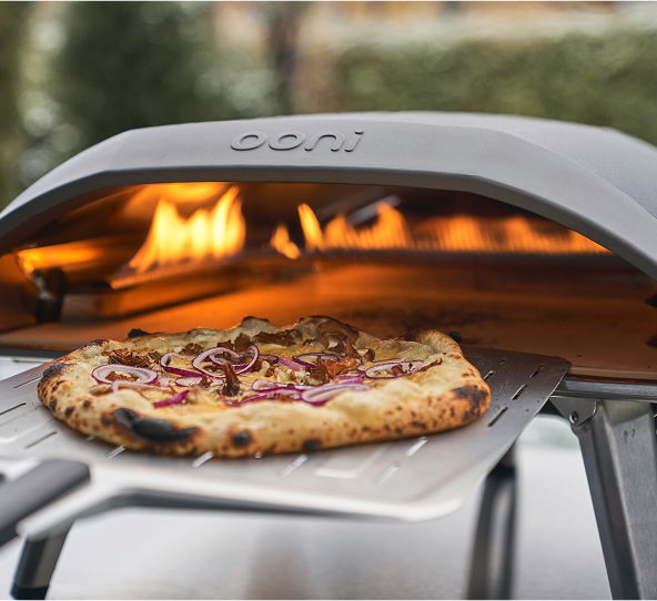 Close up glamour shot of an ooni pizza oven with a pizza on the perforated pizza peel bringing out a crisp pizza
