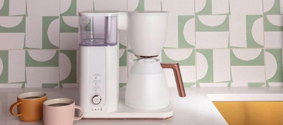 A zoomed-in view of a white and copper cafe coffee maker on a kitchen counter. The background has a soft green and white patterned backsplash