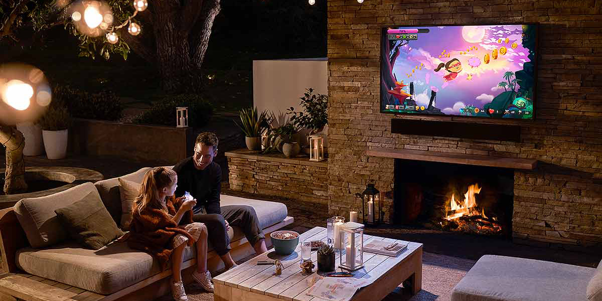 A father and daughter in their outdoor patio sofa space watching an outdoor TV mounted to an outdoor fireplace