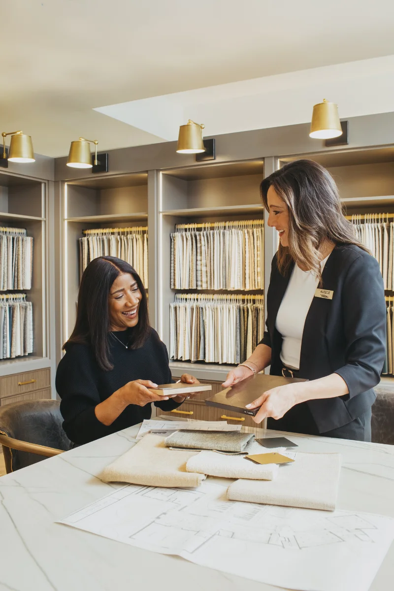 Two people sit at a table with design books, samples, and drawings of furniture