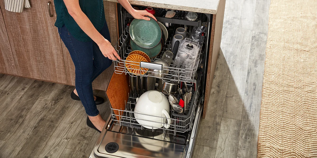 A woman adding a plate to an open, almost full dishwasher