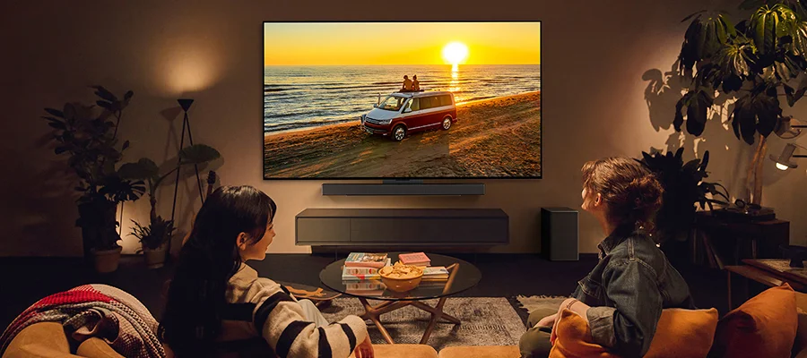 Two people sitting on the couch watching TV. The TV displays an image of two people sitting on top of a van watching the sunset by the beach.