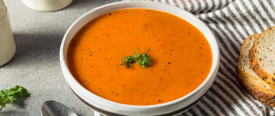 A bowl of roasted red pepper soup with a sprig of herbs sitting next to some bread and a spoon