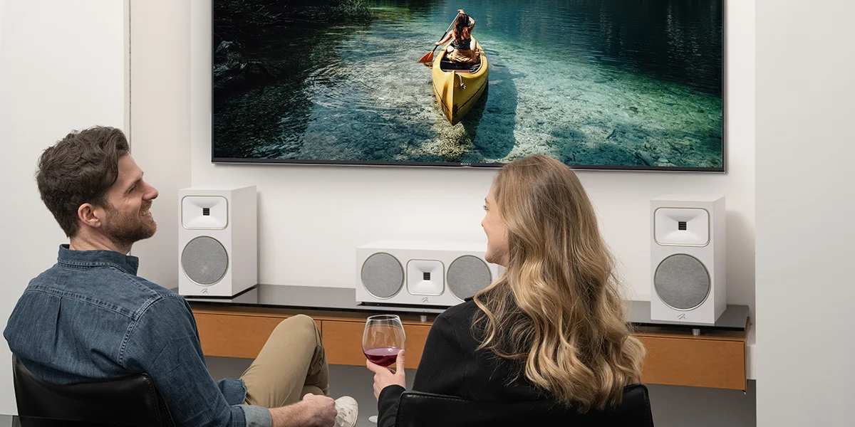 A man and a woman watching TV in a living room with three white bookshelf speakers on the TV stand