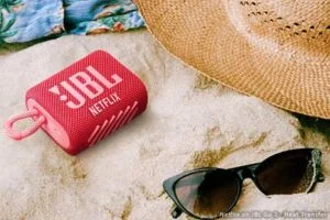 Pink JBL portable speaker on sand next to sunglasses and a straw hat at the beach.