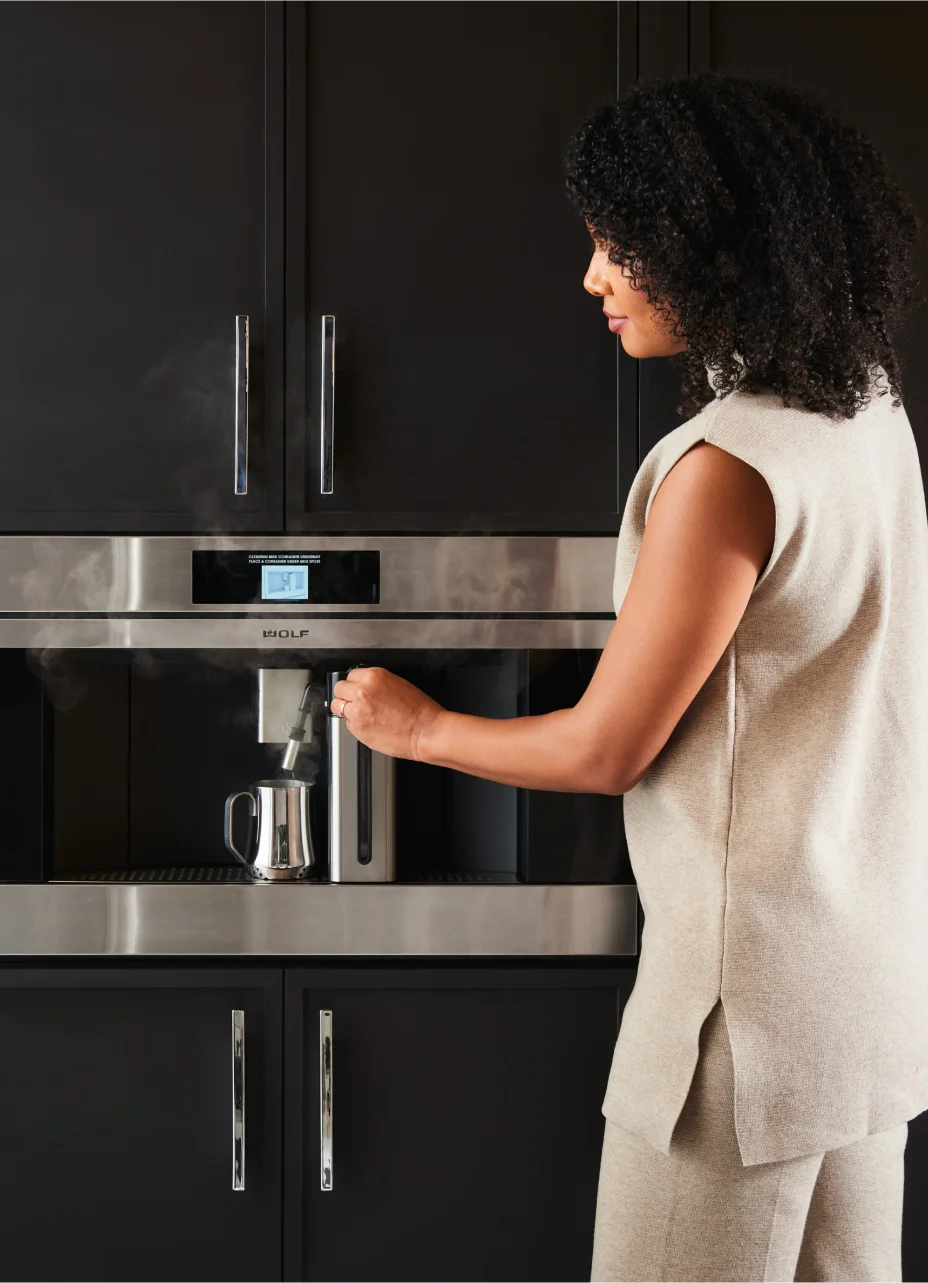 A woman operates a Wolf built-in coffee machine in a modern kitchen, preparing a fresh cup of coffee