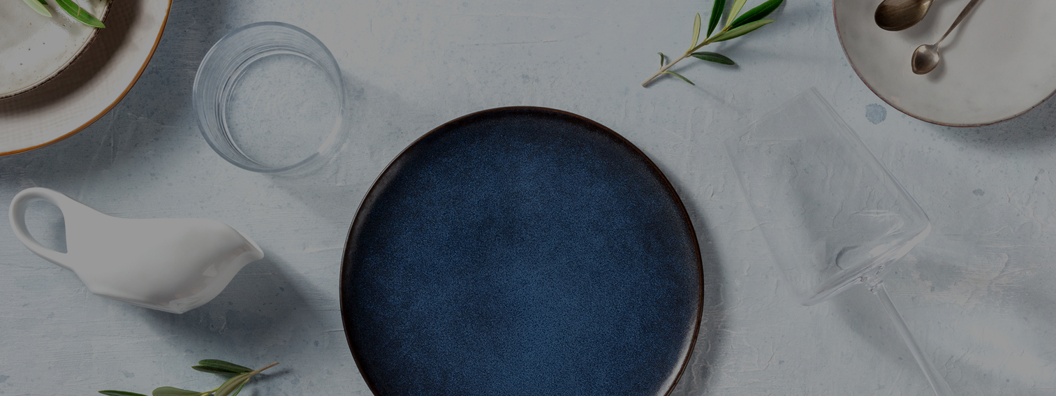An overhead shot of a blue plate with cutlery and glassware and serve-ware situated together showing a table ready for a meal