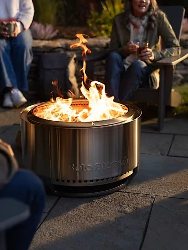 Tight shot on a Solo Stove firepit lit outdoors with people gathering around with warm beverages
