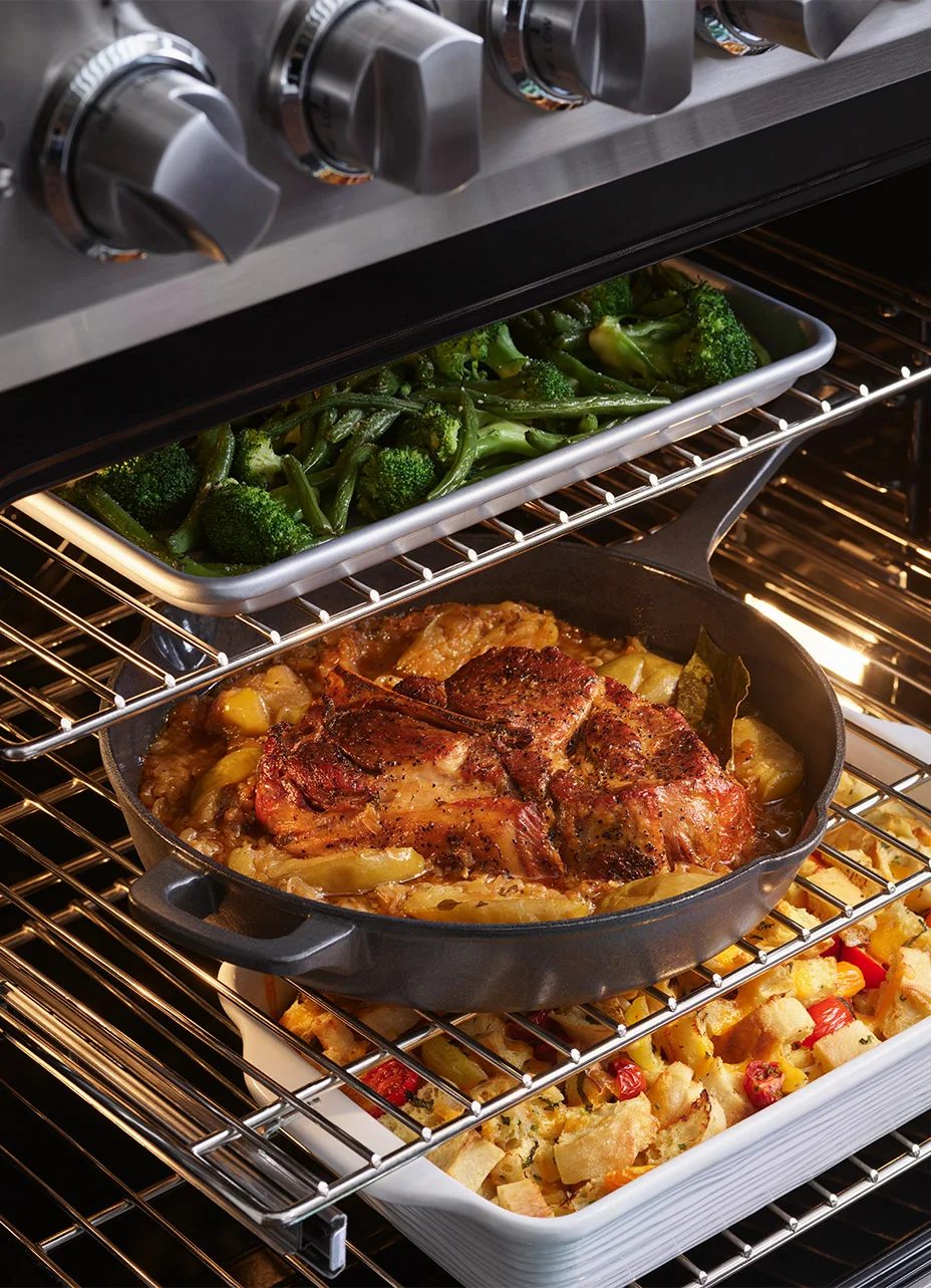 a close up on the inside of a viking range showcasing multiple cookware products with broccoli cooking in one, beef cooking in the second shelf and another roasting pan with food cooking as well