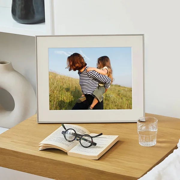 An Aura Walden digital photo frame sitting on a wooden table next to a book and a glass of water displaying a photo of a woman giving a child a piggy back ride