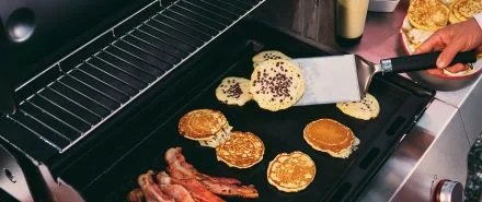 Breakfast foods like pancakes and bacon being cooked and flipped on a Weber griddle
