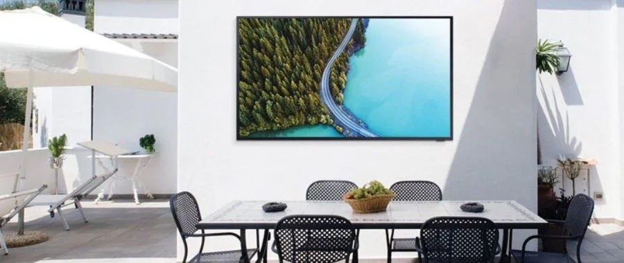 A sunny scene with an outdoor TV up against a wall with a set of patio furniture'