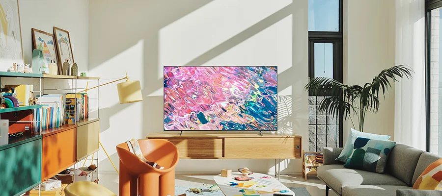 The Samsung Q60D TV sitting on a wooden stand in a color living room. The TV is displaying pink and blue colors.