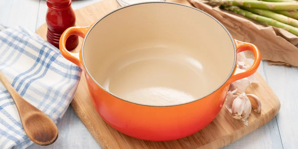A clean, orange Dutch oven sitting on a cutting board next to some garlic and asparagus.