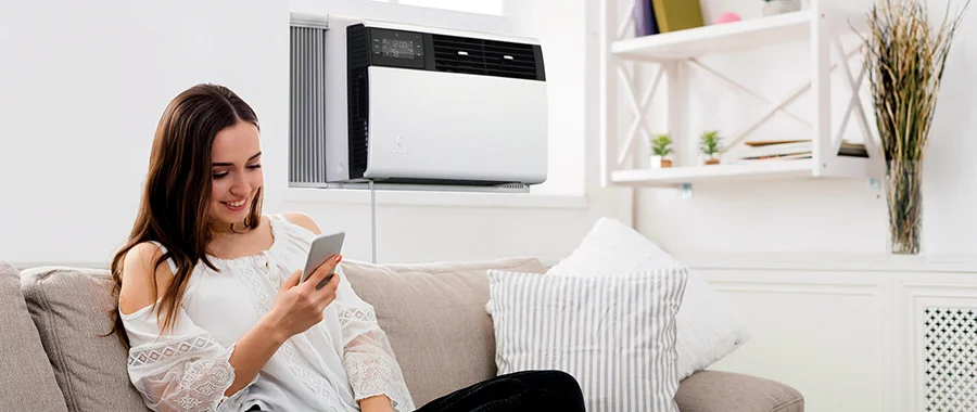 A woman scrolling on her smartphone with a window air conditioner unit installed behind her