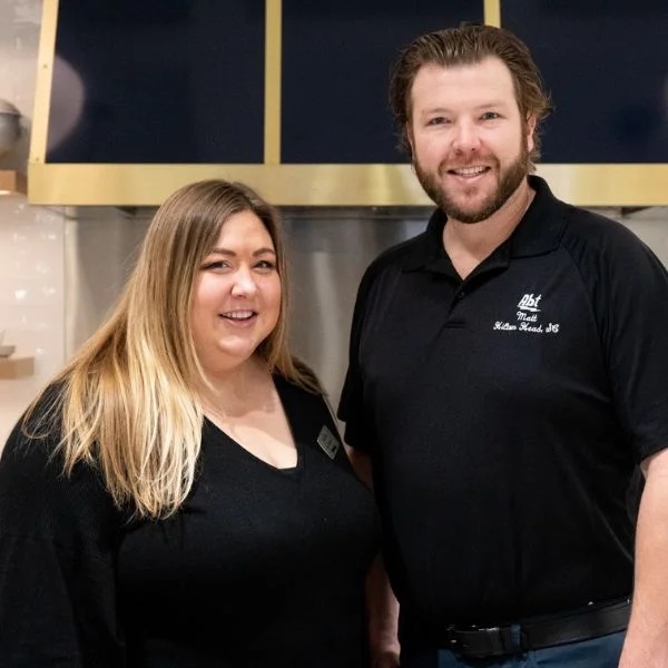 Maura and Matt Moser in front of a gold and black range hood in a model kitchen