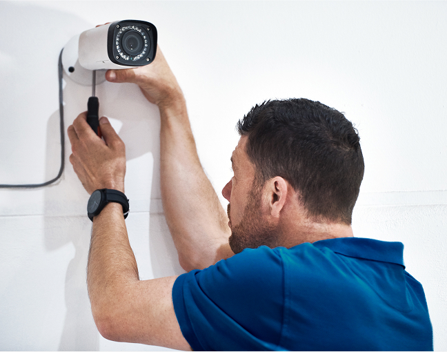 Technician in blue shirt installing a security camera on a white wall using a screwdriver.