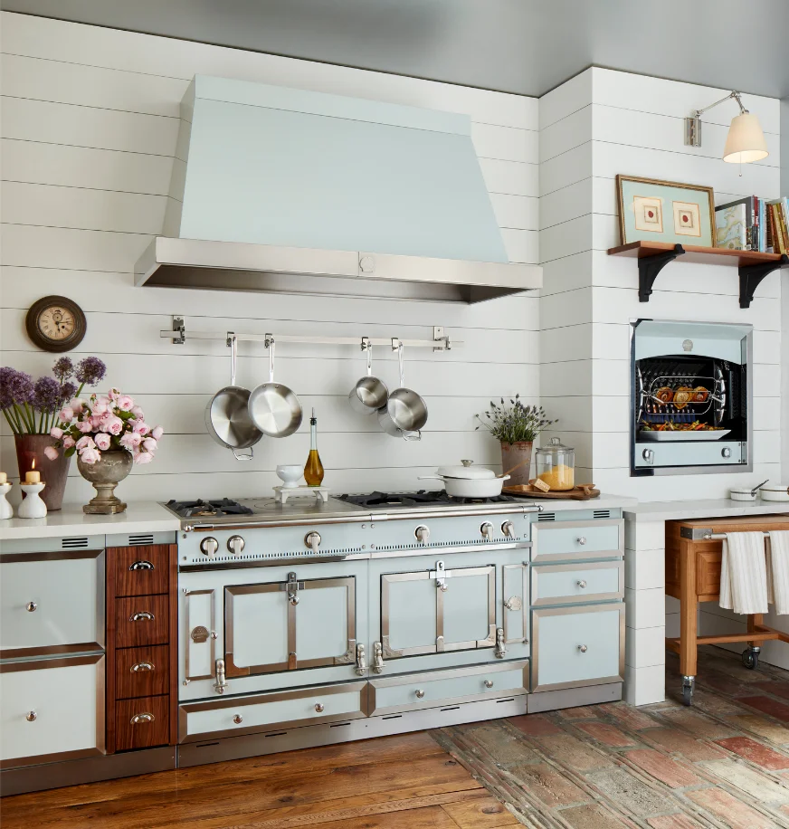 Pale blue-gray La Cornue range in a shiplap kitchen with a hanging pot rack and built-in rotisserie