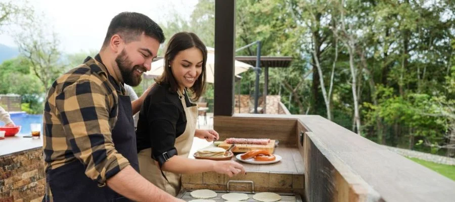 A couple grilling tortillas, meats and other foods in their outdoor kitchen. They're both wearing aprons.