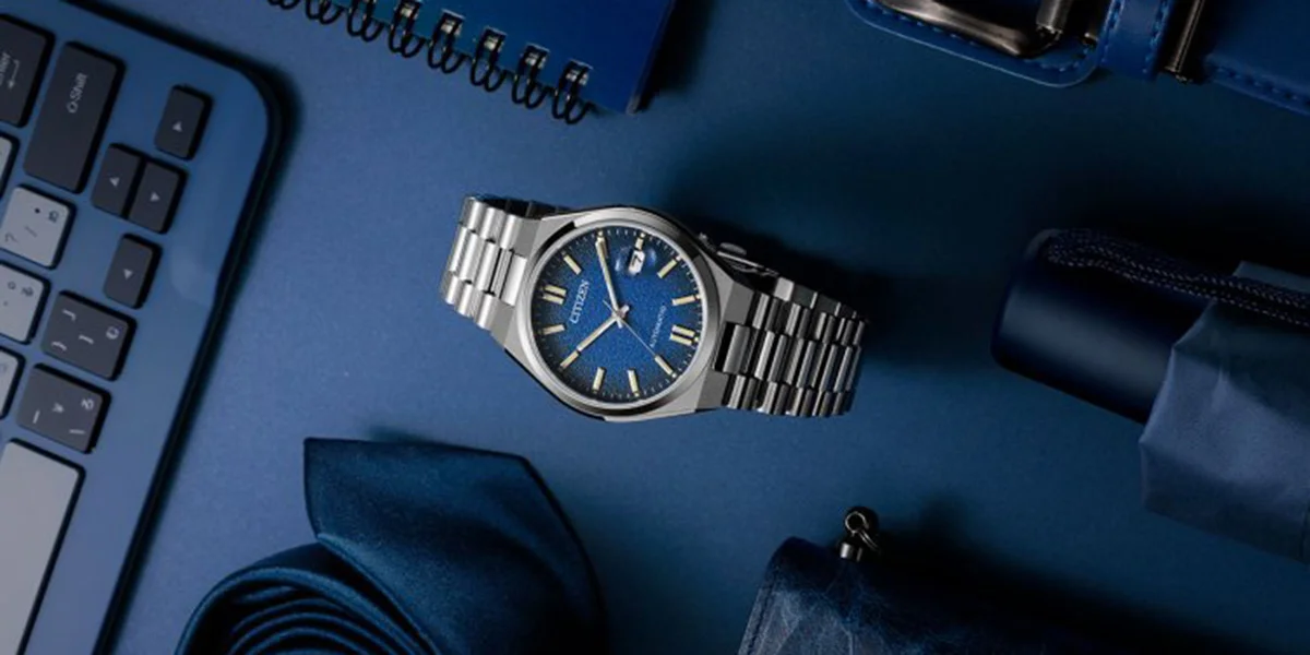 A Citizen watch with a blue face on a blue table beside accessories like a notebook, umbrella, keyboard and more