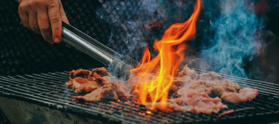 Someone grilling meat over hot flames on the grates of an outdoor grill