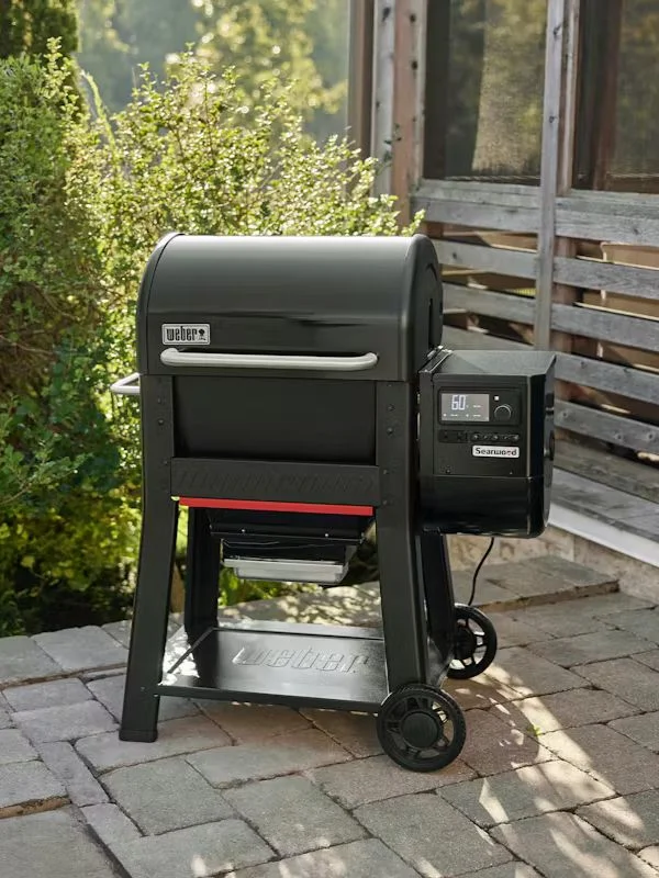A black Weber wood pellet grill on a stoned backyardwith shurbs and a wooden deck off in the back